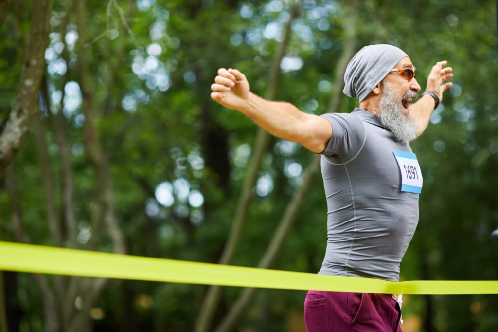 An older man with full grey beard, arms outstretched as he crosses a finishing line with a look of achievement on his face.