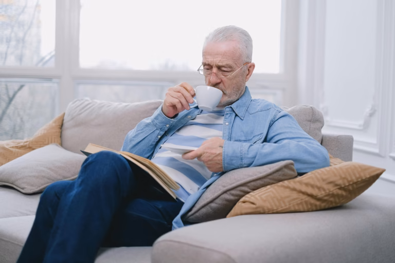 Older man relaxing at home on a sofa reading with a cup of coffee, representing a thoughtful and informed approach to PEMF therapy for seniors